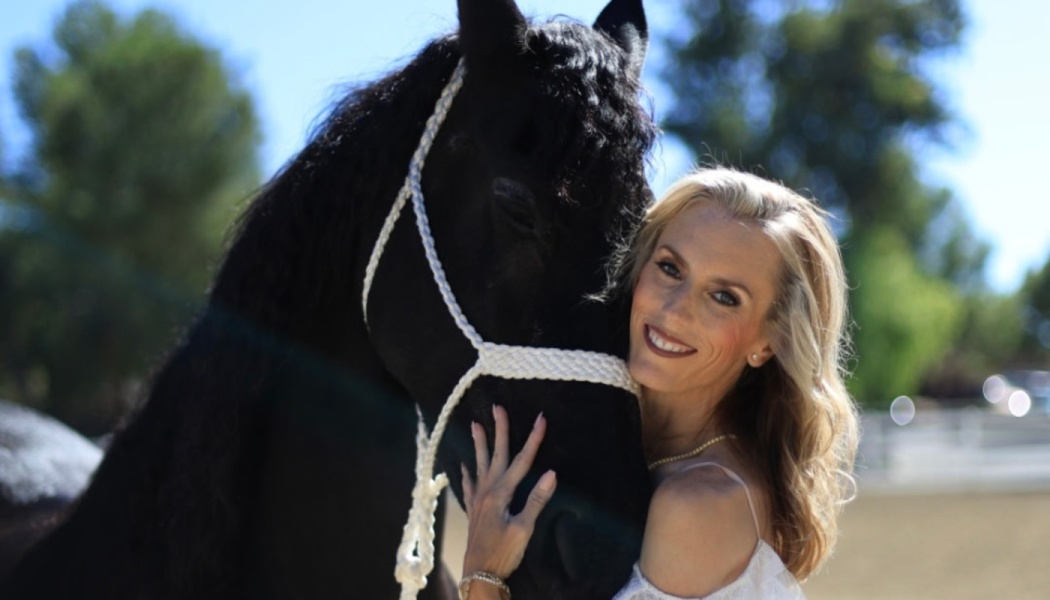 A smiling blonde woman affectionately hugging a large black horse wearing a white halter outdoors.
