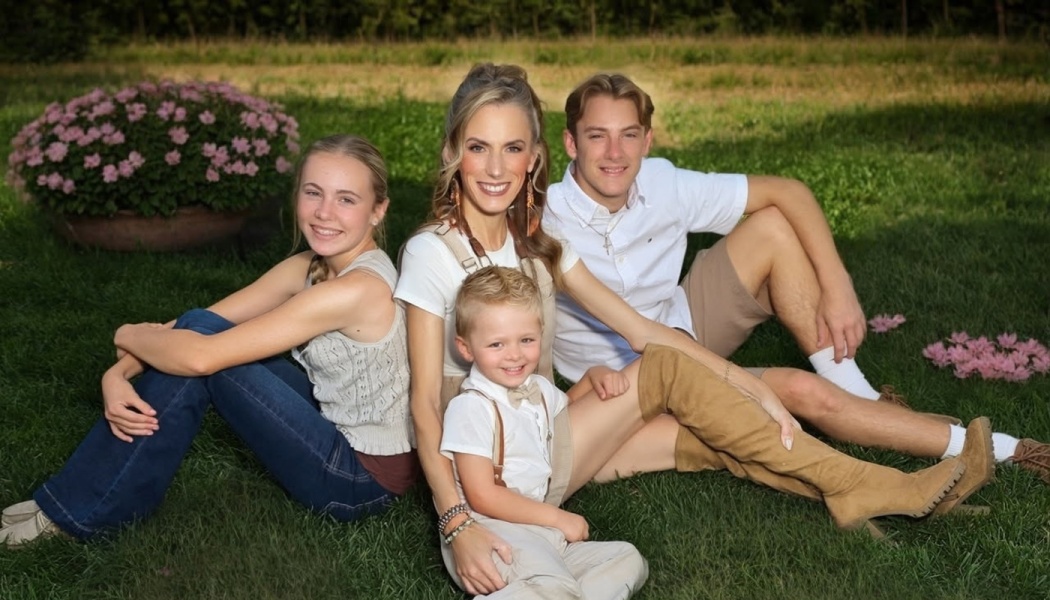 A smiling mother sitting on a grassy lawn with her teenage daughter, teenage son, and young boy.