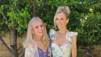 Two smiling blonde women posing together outdoors in front of lush green trees.
