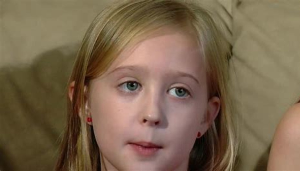 Young blonde girl with blue eyes and red earrings looking off-camera with a somber expression.