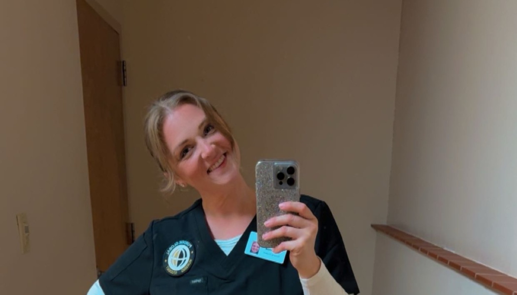 A smiling woman in black medical scrubs and an ID badge takes a mirror selfie with her smartphone.