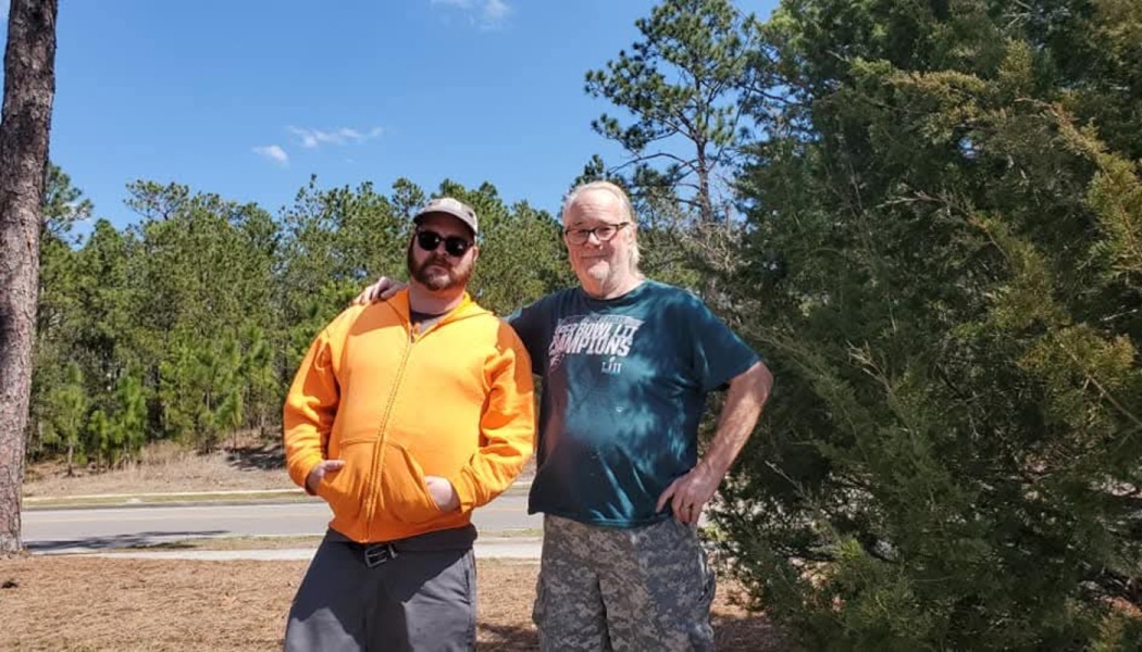 Two men pose outdoors, one wearing a bright orange hoodie and cap, the older man with an arm around him.