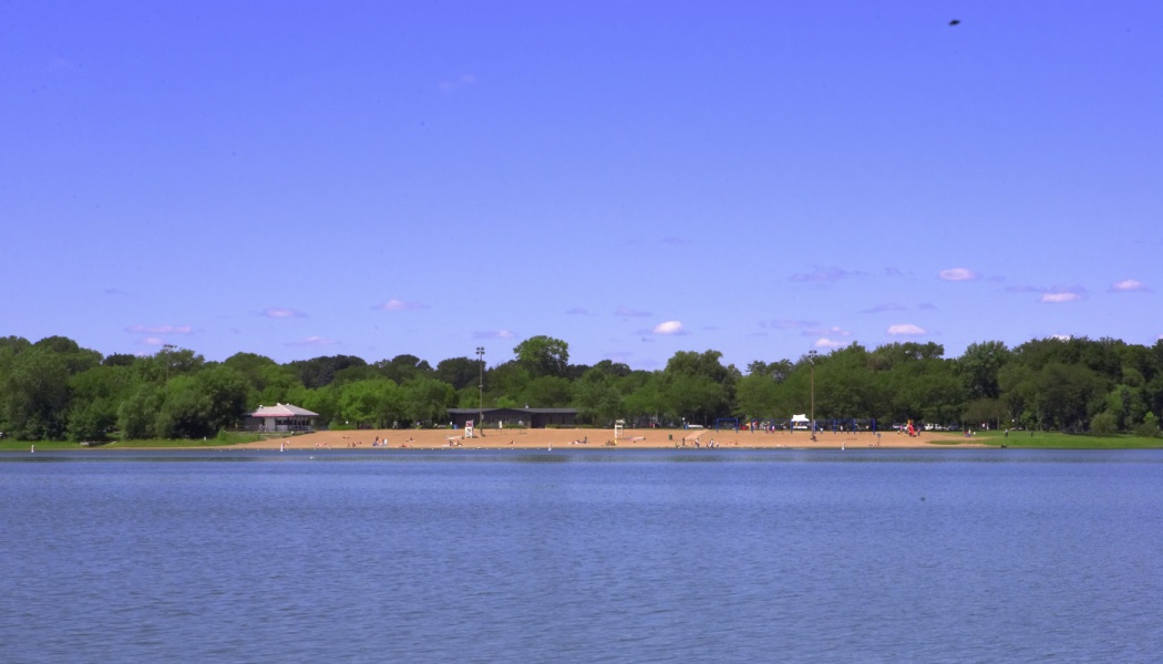 A calm blue lake with a sandy beach and lush green trees along the distant shore under a clear sky.