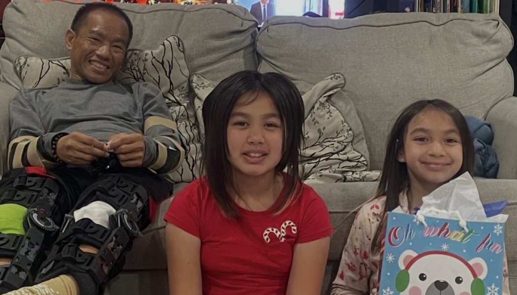 A smiling man wearing leg braces sits on a sofa behind two young girls, one holding a holiday gift bag.