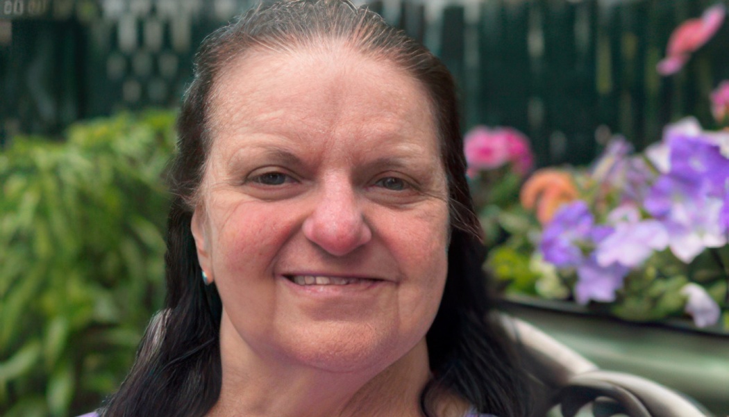 Close-up of a smiling woman with dark hair in a garden with colorful flowers in the background.