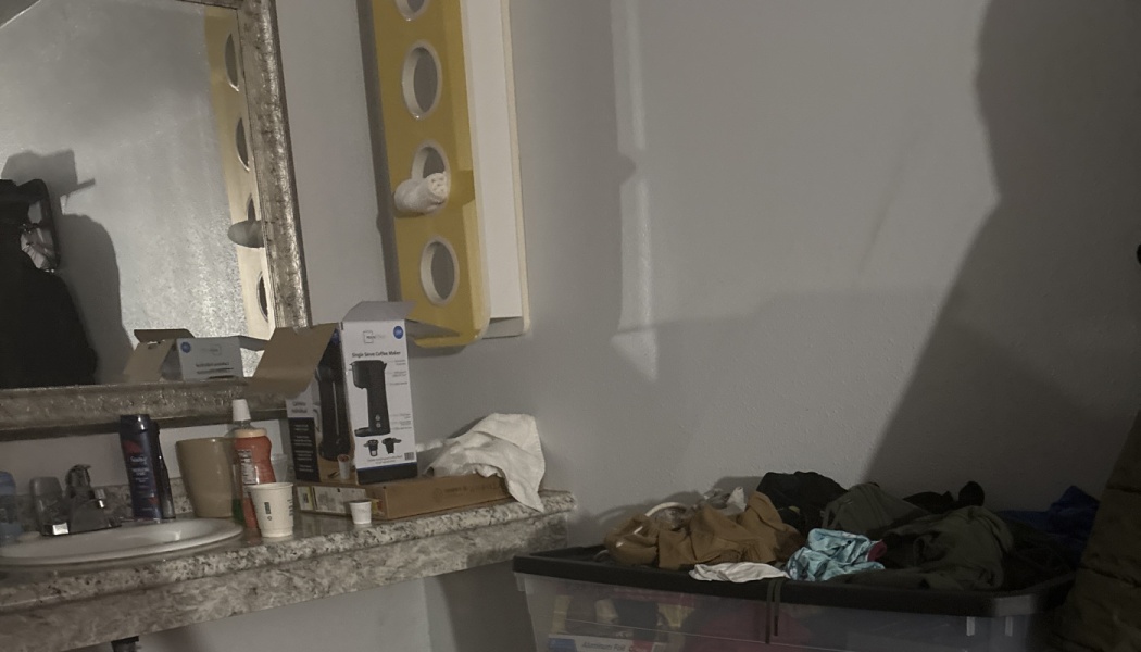 Cluttered bathroom counter with toiletries, a coffee maker box, and a storage tub filled with clothes.