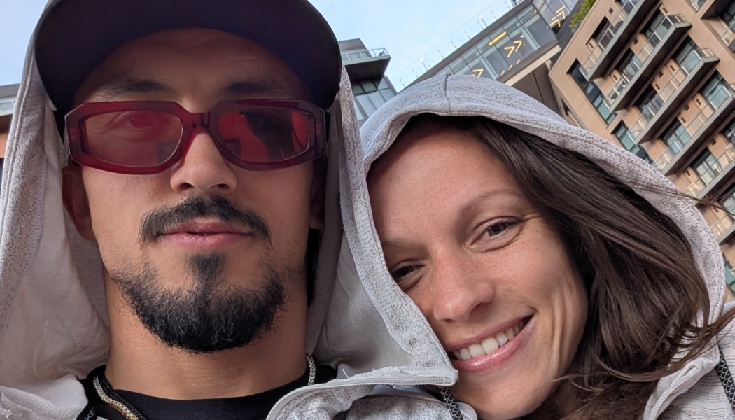 Close-up selfie of a smiling couple in grey hoodies, with the man wearing red-tinted sunglasses.