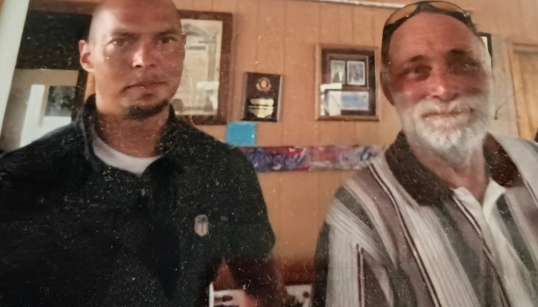 A bald younger man with a goatee and a smiling older man with a white beard pose together indoors.