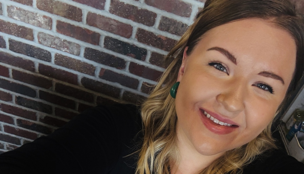 A smiling woman with blonde hair and green earrings poses in front of an exposed brick wall.