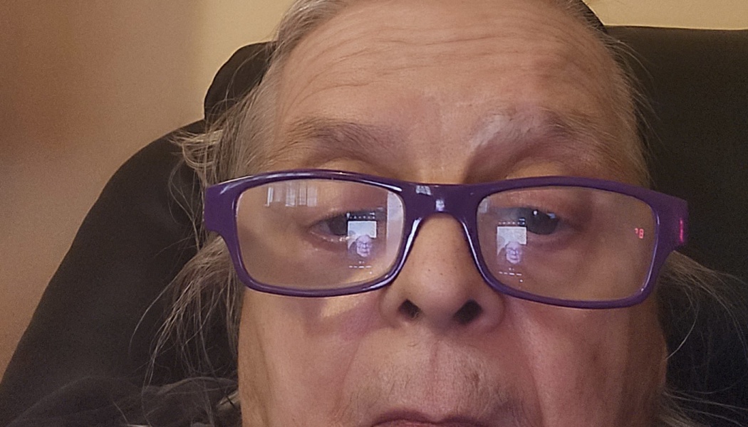 Close-up of an older person wearing purple glasses with a phone reflection in the lenses.