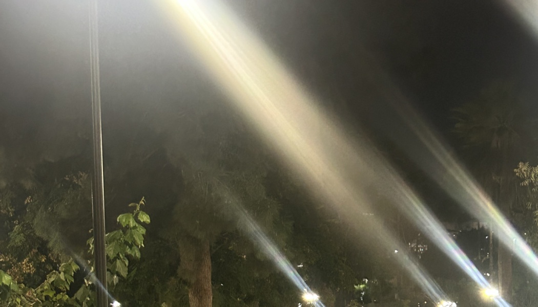 Bright light beams cutting diagonally through misty night air above dark trees.