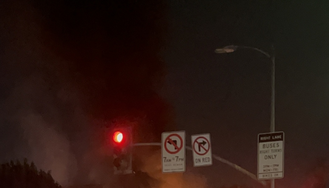 Red traffic light and street signs at night with thick smoke billowing in the background.