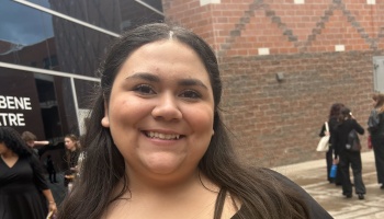 A smiling young woman with long dark hair poses outdoors in front of a brick building.