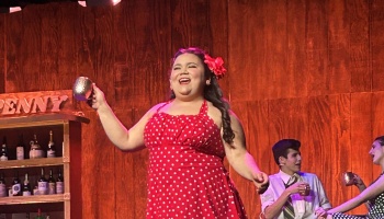 A woman in a red polka-dot dress with a flower in her hair sings joyfully on stage holding a mug.