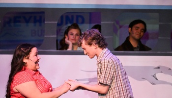 Two actors hold hands and smile on stage in front of a Greyhound bus set with passengers.