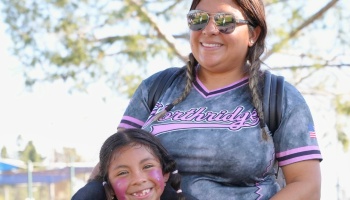 A smiling woman in aviators and a sports jersey poses outdoors with a young girl wearing pink face paint.
