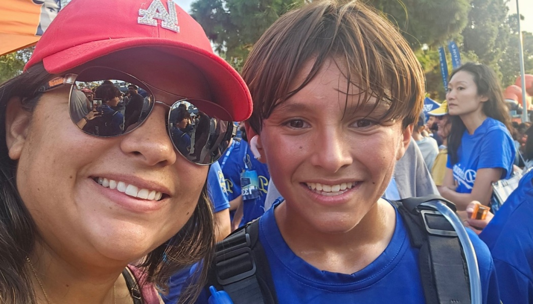 A smiling woman in a red baseball cap and sunglasses takes a selfie with a boy in a blue athletic shirt outdoors.