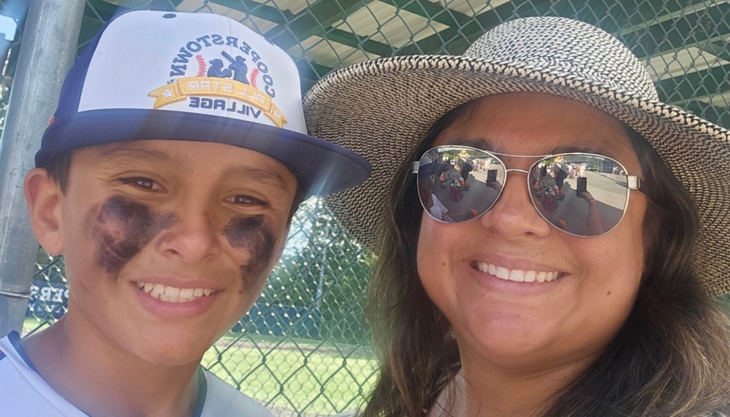 Smiling young baseball player with eye black and a woman wearing a straw hat and sunglasses at a baseball field.