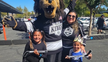 A mother and two young daughters holding mini hockey sticks pose with the LA Kings lion mascot outdoors.
