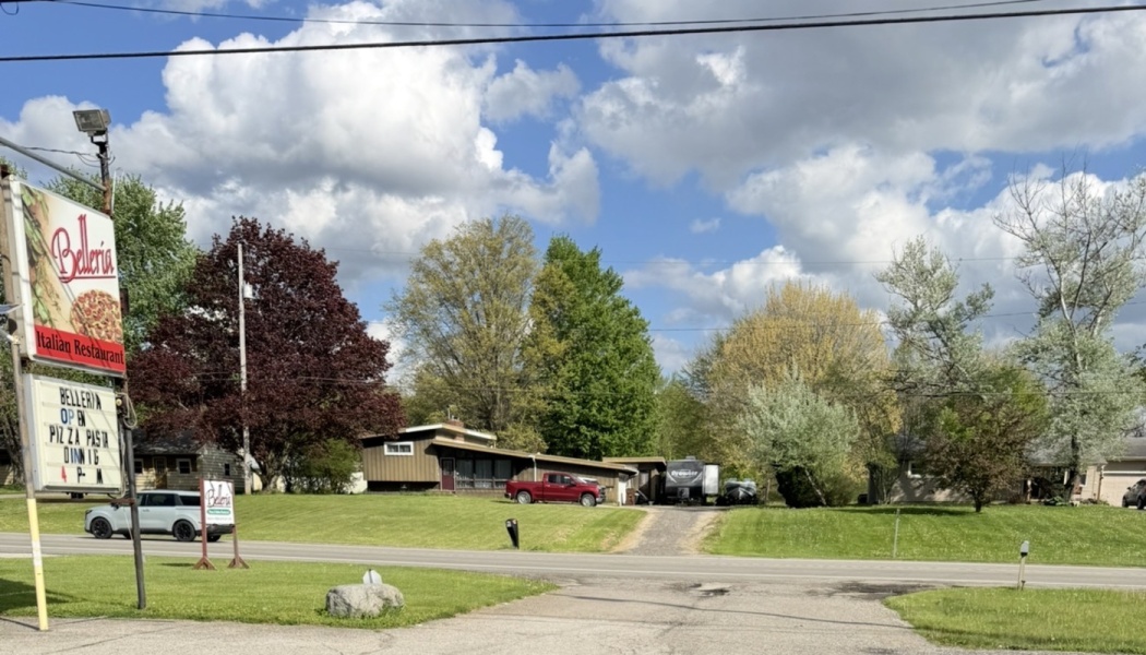 Roadside sign for Belleria Italian Restaurant with houses, green lawns, and lush trees under a partly cloudy sky.