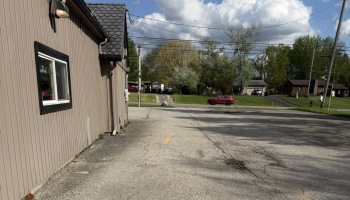 Tan metal building alongside a cracked asphalt lot leading to a residential street with trees and houses.