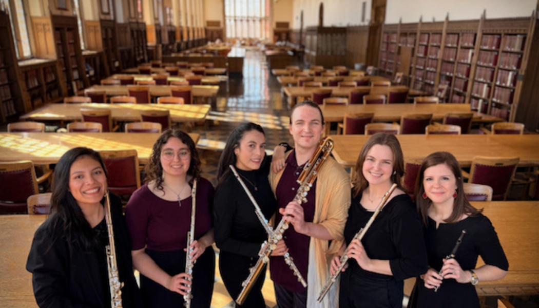 Six smiling musicians holding various flutes pose together inside a large, historic library.