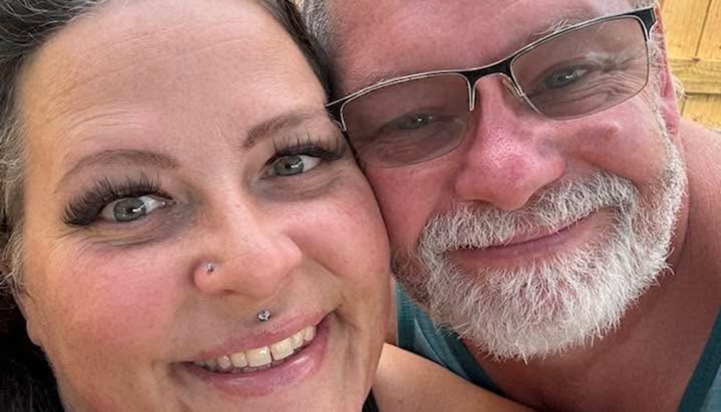 Close-up selfie of a smiling woman with facial piercings and a man with glasses and a gray beard.