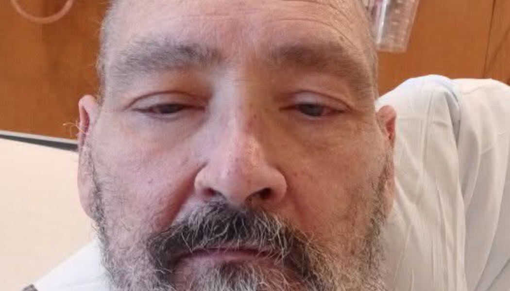 Close-up of a bearded man with tired eyes wearing a hospital gown.