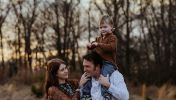 A young family outdoors with a toddler riding on his father's shoulders beside his smiling mother.