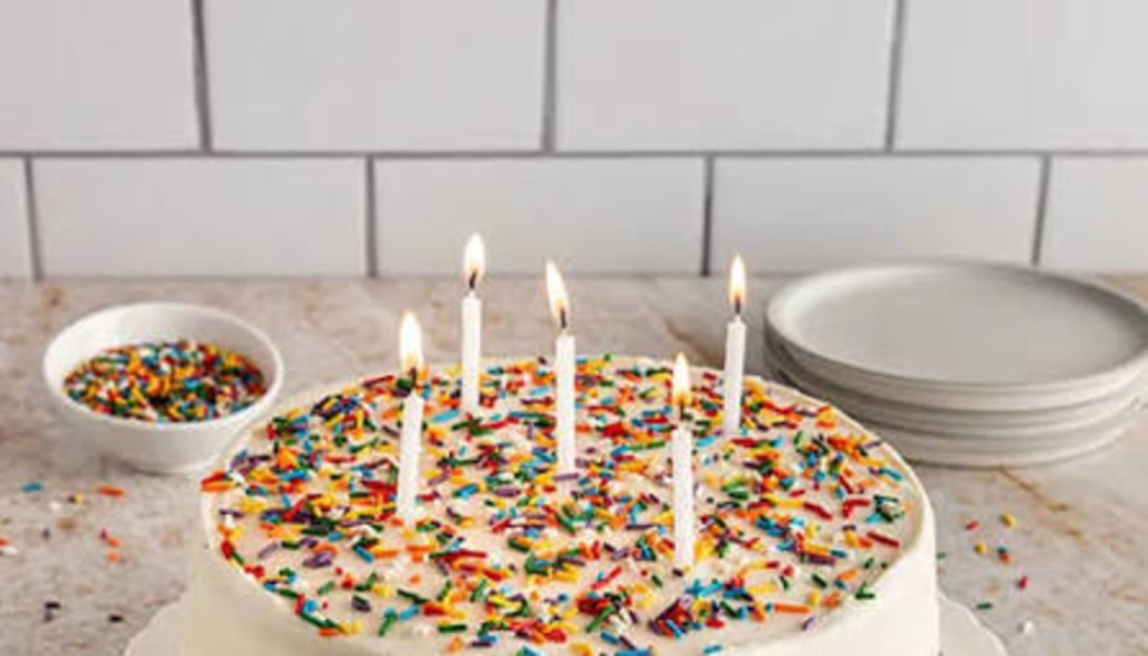 White frosted birthday cake covered with colorful sprinkles and topped with five lit white candles.