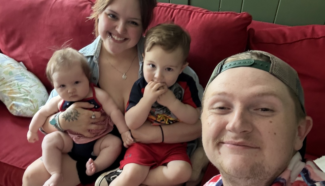 A smiling young family of four with a baby and toddler sitting together on a red couch.