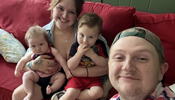 A smiling young family of four with a baby and toddler sitting together on a red couch.