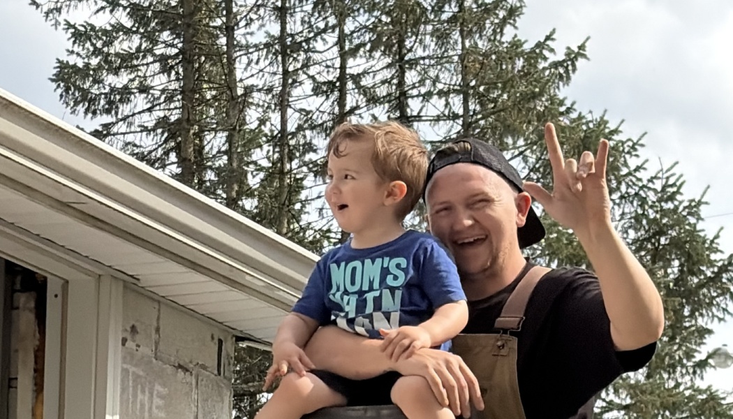 A smiling man in overalls holds a toddler on his shoulders while throwing a rock-on sign outdoors.