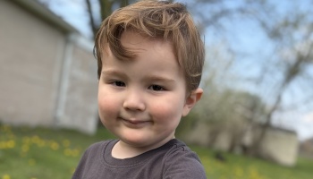 A young boy with light brown hair smiling gently at the camera outdoors.
