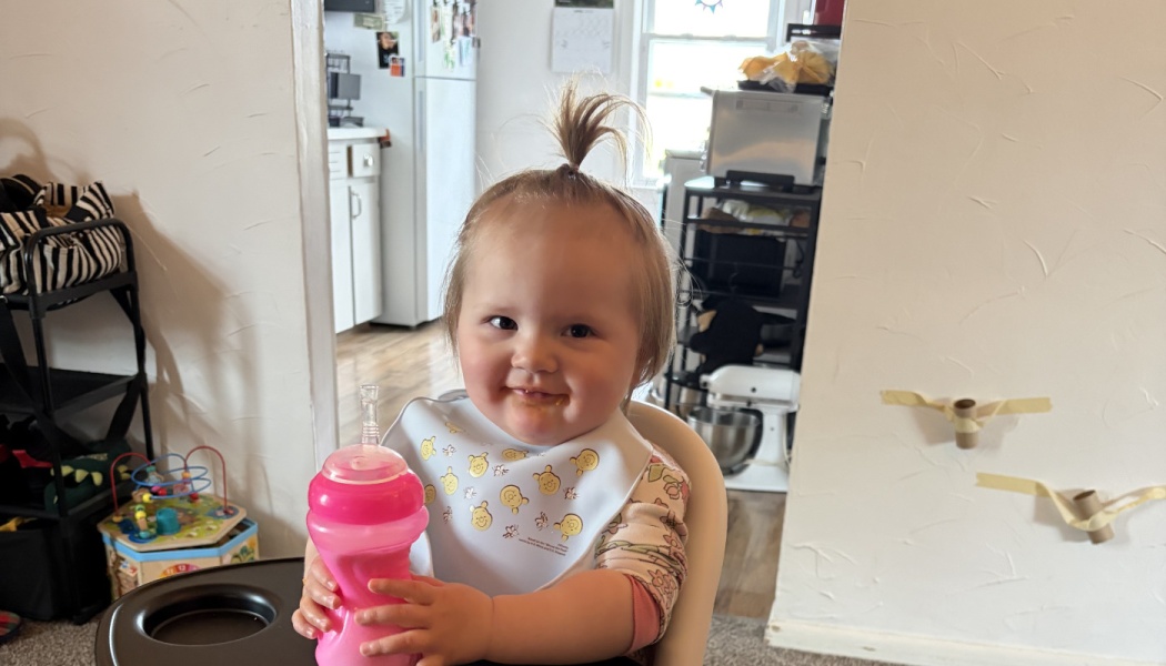 Smiling toddler with a topknot ponytail sitting in a high chair holding a pink sippy cup.