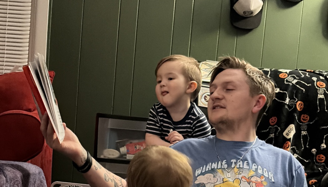 A father reads a storybook aloud to his young toddler sitting next to him.