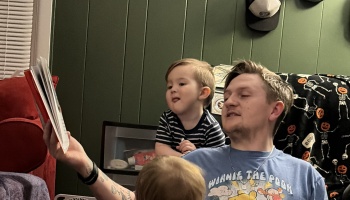 A father reads a storybook aloud to his young toddler sitting next to him.