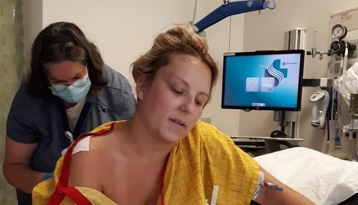 A woman in a yellow hospital gown receives care from a masked nurse in a hospital room.