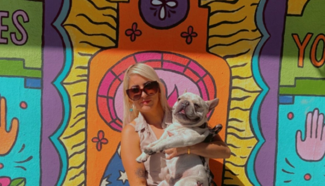 A blonde woman with sunglasses holds a French bulldog in front of a colorful painted mural.