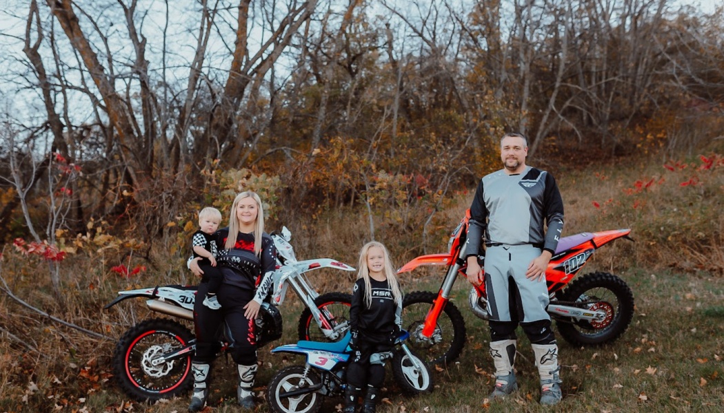A family of four in motocross gear poses outdoors with three dirt bikes in front of autumn trees.