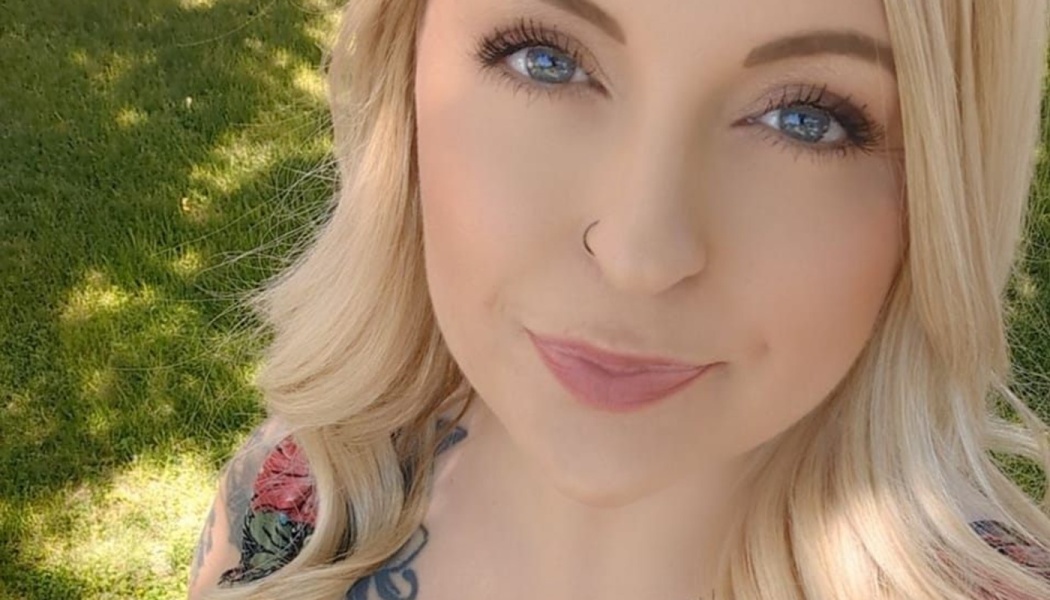 Close-up portrait of a blonde woman with blue eyes, a nose ring, and tattoos smiling outdoors.