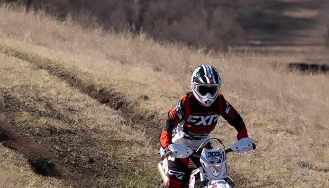 A motocross rider in red and black gear rides a white dirt bike along an off-road trail on a grassy hill.
