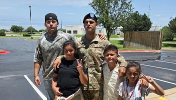 A U.S. Army soldier poses outdoors in uniform with another man and three children.