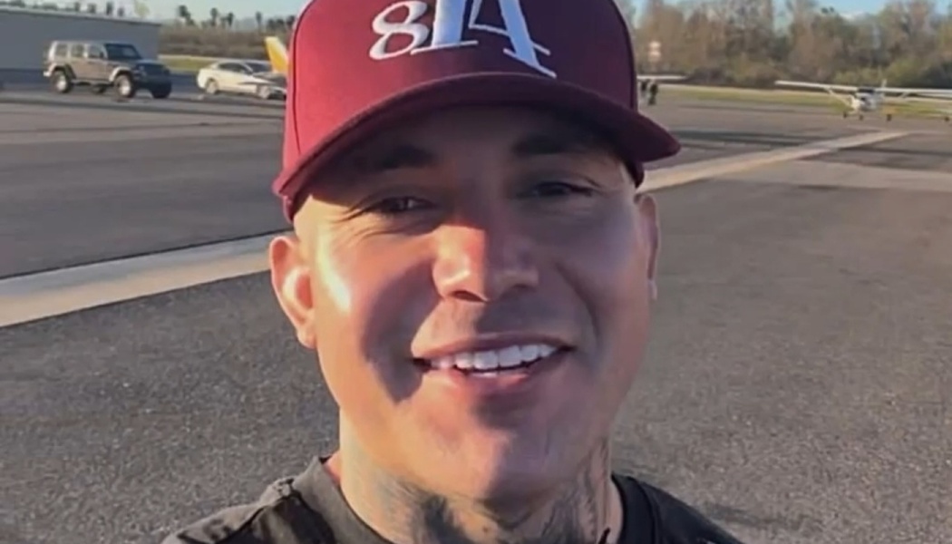 Smiling man with neck tattoos wearing a maroon baseball cap at an airfield with planes in the background.
