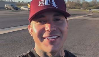 Smiling man with neck tattoos wearing a maroon baseball cap at an airfield with planes in the background.