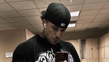 Man wearing a black baseball cap, earbuds, and graphic t-shirt taking a mirror selfie in a locker room.