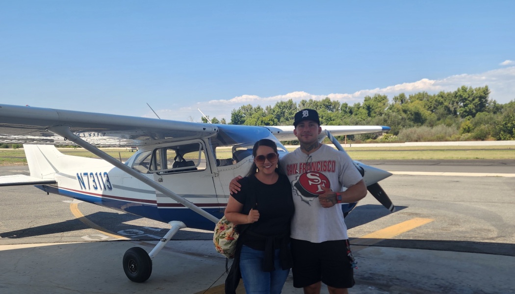 A smiling man and woman pose in front of a small white and blue propeller airplane on a sunny tarmac.