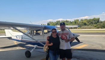 A smiling man and woman pose in front of a small white and blue propeller airplane on a sunny tarmac.