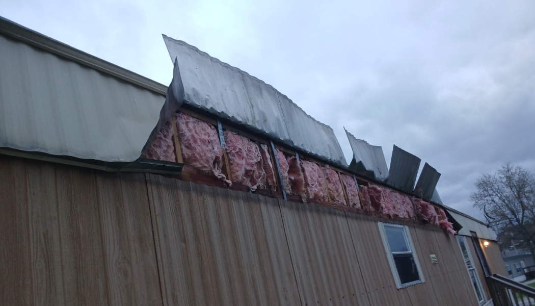 Storm damage to a mobile home roof with metal siding peeled back, exposing pink fiberglass insulation.