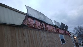 Storm damage to a mobile home roof with metal siding peeled back, exposing pink fiberglass insulation.